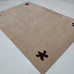 May include: A rectangular beige rug with four dark brown star-shaped designs. The rug is made of a soft material and is placed on a white tiled floor. The rug's dimensions are not visible.