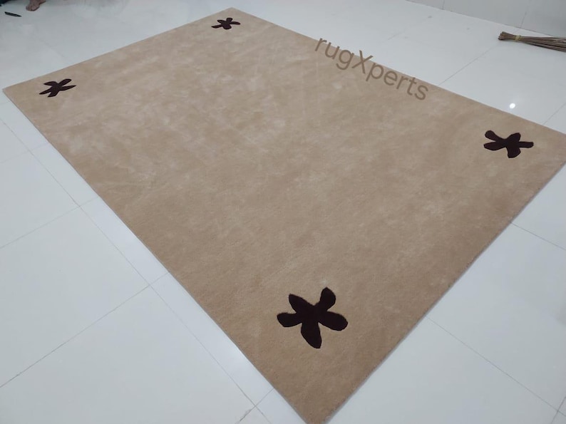 May include: A rectangular beige rug with four dark brown star-shaped designs. The rug is made of a soft material and is placed on a white tiled floor. The rug's dimensions are not visible.