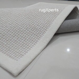 May include: A close-up of a white rug with a subtle textured pattern. The rug has a white border and is folded to show the underside, which is a light gray color. The text "rugXperts" is visible in the upper right corner of the image.