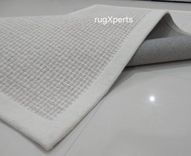 May include: A close-up of a white rug with a subtle textured pattern. The rug has a white border and is folded to show the underside, which is a light gray color. The text "rugXperts" is visible in the upper right corner of the image.