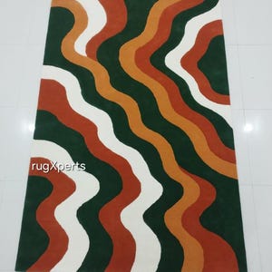 May include: A rectangular rug with a wavy pattern in shades of green, white, orange, and rust. The rug features the text "rugXperts" in the lower left corner. The design is modern and abstract.