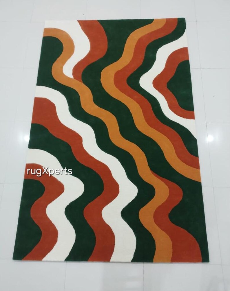 May include: A rectangular rug with a wavy pattern in shades of green, white, orange, and rust. The rug features the text "rugXperts" in the lower left corner. The design is modern and abstract.
