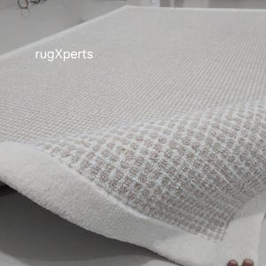 May include: A white and cream colored rug with a diamond pattern. The rug has a thick, fluffy texture and a white border.