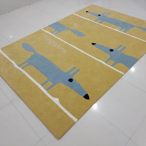 May include: A yellow rug with a repeating pattern of blue stylized foxes. The rug has white stripes running vertically across the surface. The text "rugXperts" is printed in white on the rug.