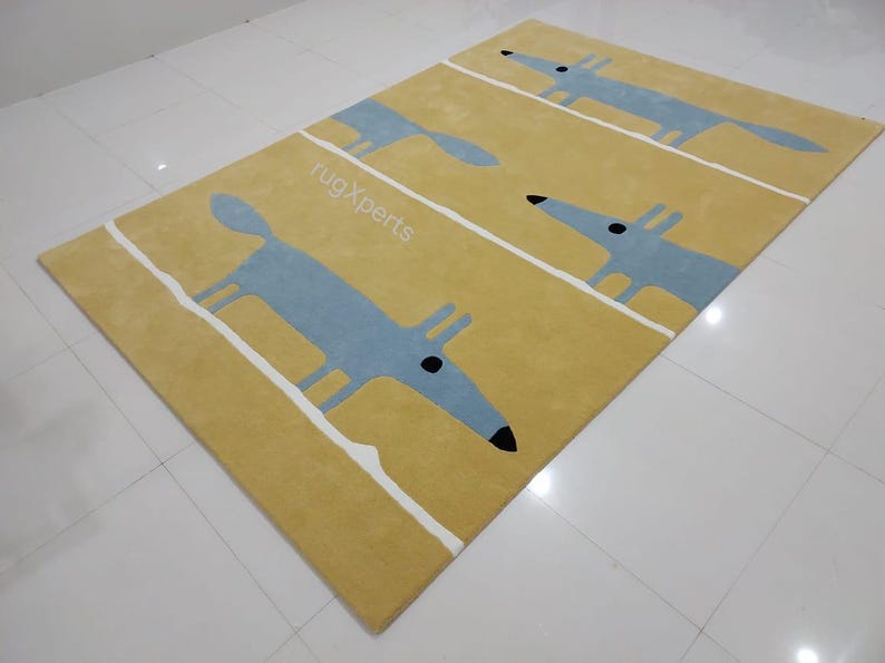 May include: A yellow rug with a repeating pattern of blue stylized foxes. The rug has white stripes running vertically across the surface. The text "rugXperts" is printed in white on the rug.