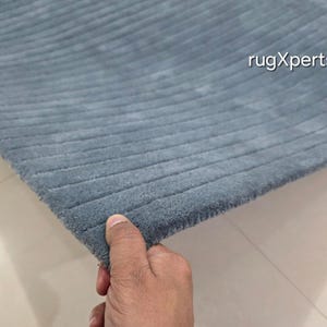 Op de afbeelding: Een gedeelte van een blauw tapijt met een geribbelde textuur. Het tapijt wordt door een hand vastgehouden, die de rand en het zachte, pluche materiaal laat zien. De tekst "rugXperts" is zichtbaar in de rechterbovenhoek.