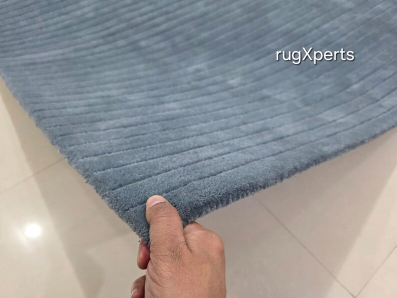 Op de afbeelding: Een gedeelte van een blauw tapijt met een geribbelde textuur. Het tapijt wordt door een hand vastgehouden, die de rand en het zachte, pluche materiaal laat zien. De tekst "rugXperts" is zichtbaar in de rechterbovenhoek.