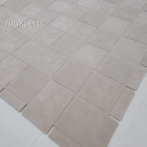 Pu&ograve; includere: Un tappeto beige chiaro composto da quadrati singoli cuciti insieme. Il tappeto &egrave; steso su una superficie bianca.