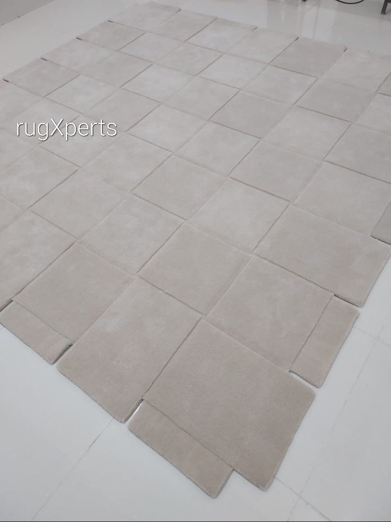 Pu&ograve; includere: Un tappeto beige chiaro composto da quadrati singoli cuciti insieme. Il tappeto &egrave; steso su una superficie bianca.