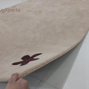 May include: Beige rectangular rug with a dark brown abstract design near the edge. The rug's texture is soft and plush, with a subtle sheen. The brand name "rugXperts" is visible in the top left corner. The rug is partially rolled up, revealing its underside.