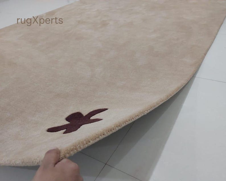 May include: Beige rectangular rug with a dark brown abstract design near the edge. The rug's texture is soft and plush, with a subtle sheen. The brand name "rugXperts" is visible in the top left corner. The rug is partially rolled up, revealing its underside.