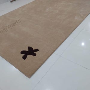 May include: A rectangular beige rug with a dark brown decorative design. The rug has a soft texture and is placed on a white tiled floor. The text "rugXperts" is visible on the rug.