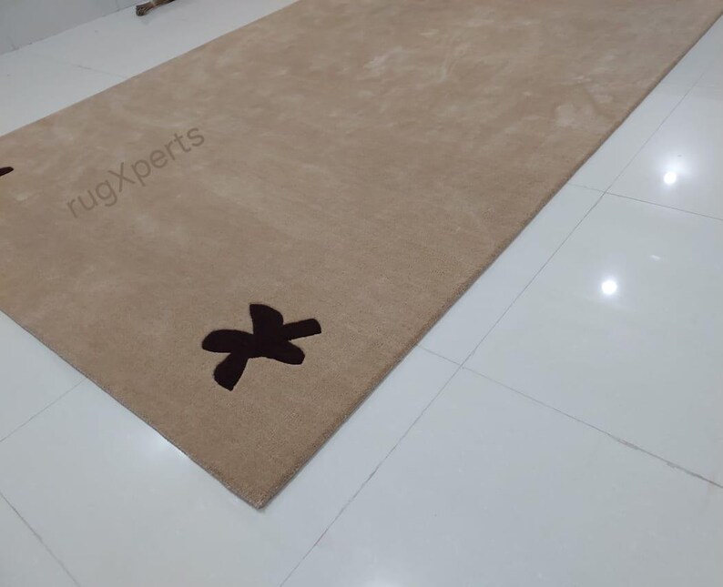 May include: A rectangular beige rug with a dark brown decorative design. The rug has a soft texture and is placed on a white tiled floor. The text "rugXperts" is visible on the rug.