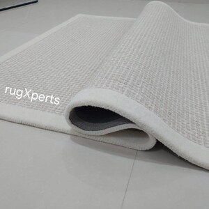 May include: A white rug with a textured surface and a white border. The rug is rolled up and the text "rugXperts" is visible.