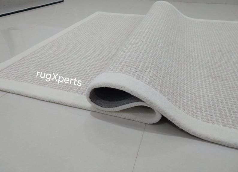 May include: A white rug with a textured surface and a white border. The rug is rolled up and the text "rugXperts" is visible.