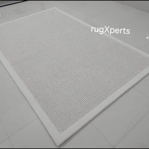 May include: A white area rug with a subtle textured pattern and a white border. The rug is on a white tiled floor. rugXperts