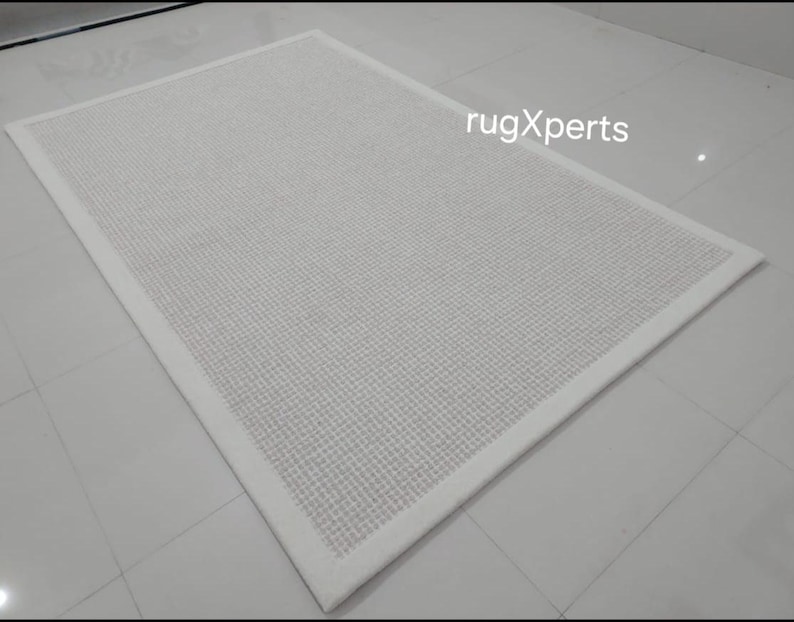 May include: A white area rug with a subtle textured pattern and a white border. The rug is on a white tiled floor. rugXperts