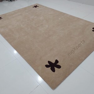 May include: A rectangular beige rug with four dark brown star-shaped designs. The rug has a soft texture and is placed on a white tiled floor. The text "rugXperts" is visible on the rug.