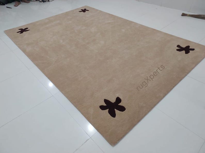 May include: A rectangular beige rug with four dark brown star-shaped designs. The rug has a soft texture and is placed on a white tiled floor. The text "rugXperts" is visible on the rug.