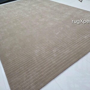 Op de afbeelding: Een groot, rechthoekig beige tapijt met een geribbelde textuur. Het tapijt is gemaakt van een zacht materiaal en heeft een subtiele glans. De tekst "rugXperts" is zichtbaar in de afbeelding. Dit tapijt is geschikt voor woonkamers en slaapkamers.