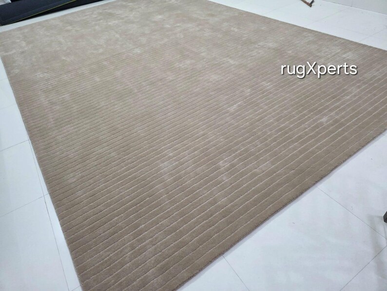 Op de afbeelding: Een groot, rechthoekig beige tapijt met een geribbelde textuur. Het tapijt is gemaakt van een zacht materiaal en heeft een subtiele glans. De tekst "rugXperts" is zichtbaar in de afbeelding. Dit tapijt is geschikt voor woonkamers en slaapkamers.