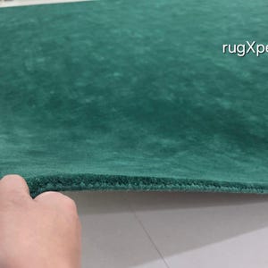 May include: A close-up of a green rug with a textured surface. The rug is a solid colour and appears to be made of a soft material. The text "rugXperts" is visible in the upper right corner.
