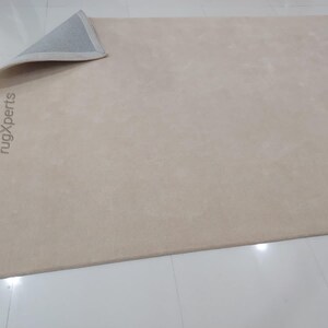 May include: A beige rectangular rug with a rolled-up corner, revealing a gray underside. The rug's surface has a subtle texture. The words "rugXperts" are printed vertically on the rug's surface.