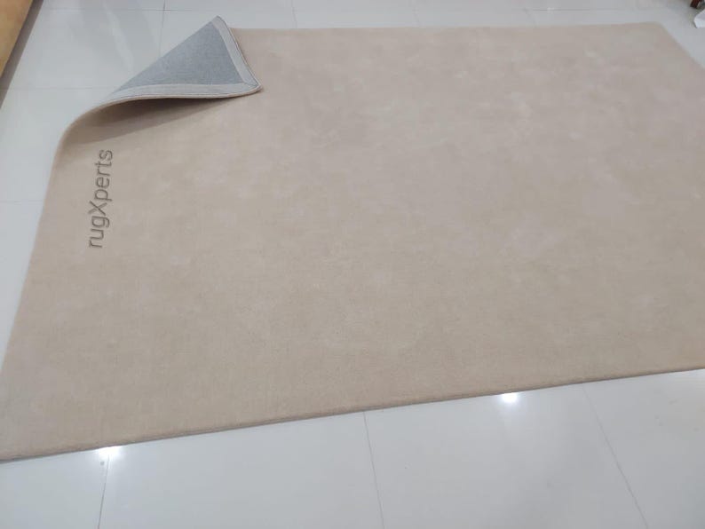 May include: A beige rectangular rug with a rolled-up corner, revealing a gray underside. The rug's surface has a subtle texture. The words "rugXperts" are printed vertically on the rug's surface.