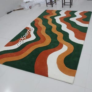 May include: A rectangular rug with a wavy pattern in green, white, orange, and rust colors. The rug has a soft texture and is suitable for living rooms or bedrooms. The text "rugXperts" is visible on the rug.