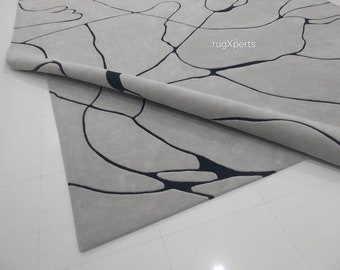Puede incluir: Primer plano de una alfombra gris y negra con un diseño abstracto. La alfombra está enrollada y el diseño es visible en la parte enrollada. La alfombra está sobre un suelo de baldosas blancas. El texto "rugXperts" es visible en la esquina superior derecha de la imagen.