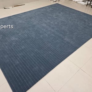 Op de afbeelding: Een groot, rechthoekig blauw tapijt met een geribbelde textuur. Het tapijt heeft een effen kleur en een subtiele glans, met de tekst "rugXperts" in de linkerbovenhoek. Het tapijt is geschikt voor een woonkamer of slaapkamer.