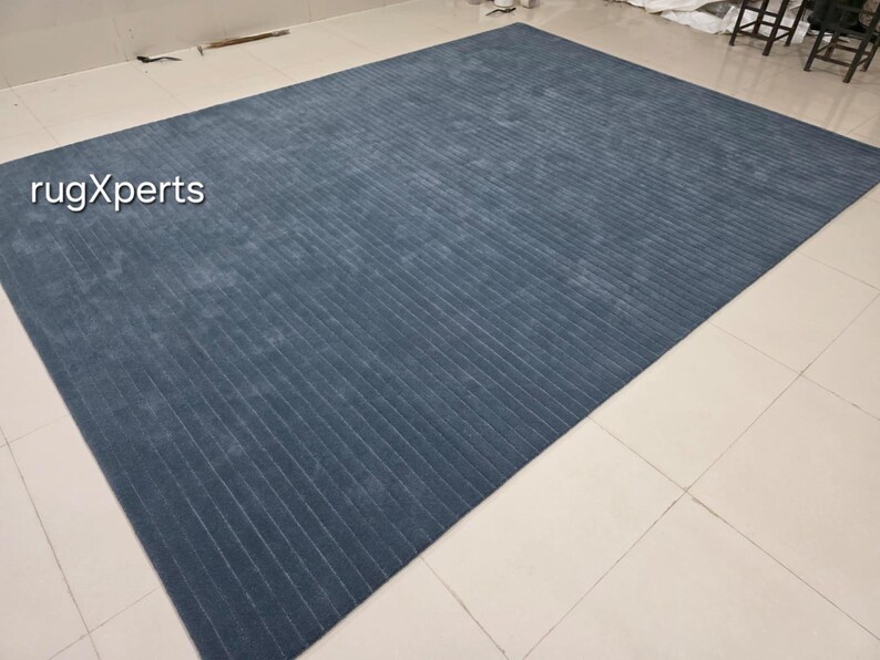 Op de afbeelding: Een groot, rechthoekig blauw tapijt met een geribbelde textuur. Het tapijt heeft een effen kleur en een subtiele glans, met de tekst "rugXperts" in de linkerbovenhoek. Het tapijt is geschikt voor een woonkamer of slaapkamer.