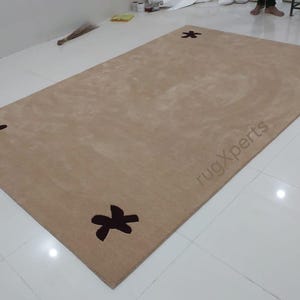 May include: A rectangular beige rug with four dark brown floral designs in each corner. The rug has a soft, plush appearance and is placed on a white tiled floor. The text "rugXperts" is visible on the rug.