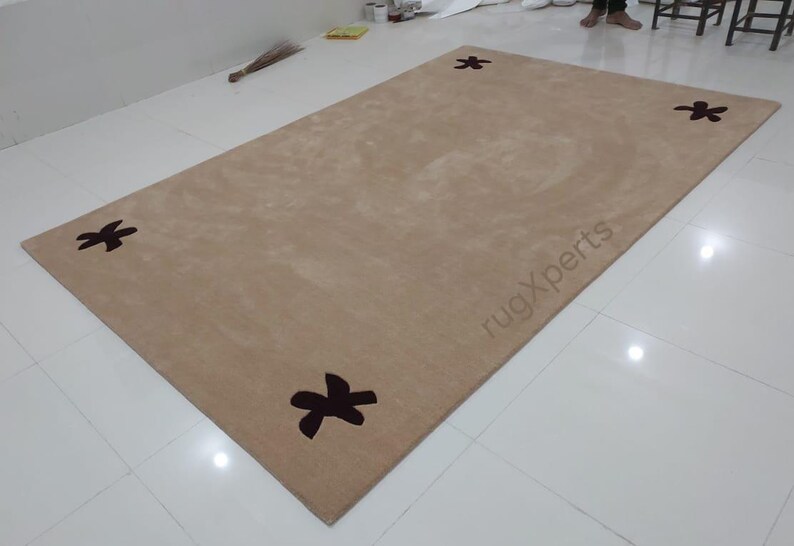 May include: A rectangular beige rug with four dark brown floral designs in each corner. The rug has a soft, plush appearance and is placed on a white tiled floor. The text "rugXperts" is visible on the rug.