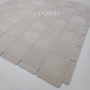 Pu&ograve; includere: Un grande tappeto quadrato di colore bianco sporco, composto da quadrati singoli cuciti insieme. Il tappeto &egrave; realizzato in un materiale morbido e soffice e presenta una texture sottile. Il tappeto &egrave; steso su un pavimento bianco. Il testo "rugXperts" &egrave; visibile al centro del tappeto.