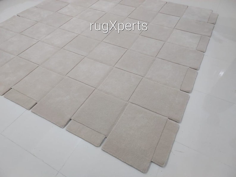 Pu&ograve; includere: Un grande tappeto quadrato di colore bianco sporco, composto da quadrati singoli cuciti insieme. Il tappeto &egrave; realizzato in un materiale morbido e soffice e presenta una texture sottile. Il tappeto &egrave; steso su un pavimento bianco. Il testo "rugXperts" &egrave; visibile al centro del tappeto.