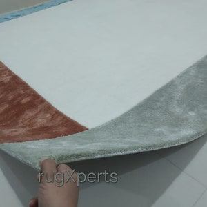 May include: A rectangular rug with a white central area and a border in shades of brown, green, and blue. The rug has a soft, plush texture, with a folded edge showing the underside. The rug is designed for home decor.