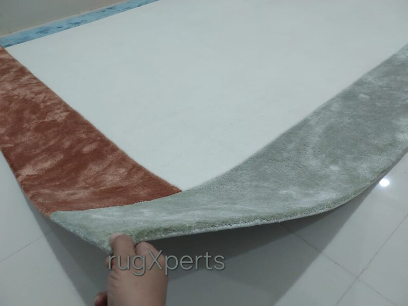 May include: A rectangular rug with a white central area and a border in shades of brown, green, and blue. The rug has a soft, plush texture, with a folded edge showing the underside. The rug is designed for home decor.