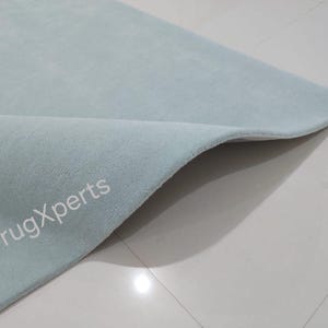 May include: A light blue rug with a soft, textured surface. The corner of the rug is folded over, revealing the underside. The text "rugXperts" is visible on the rug's surface.
