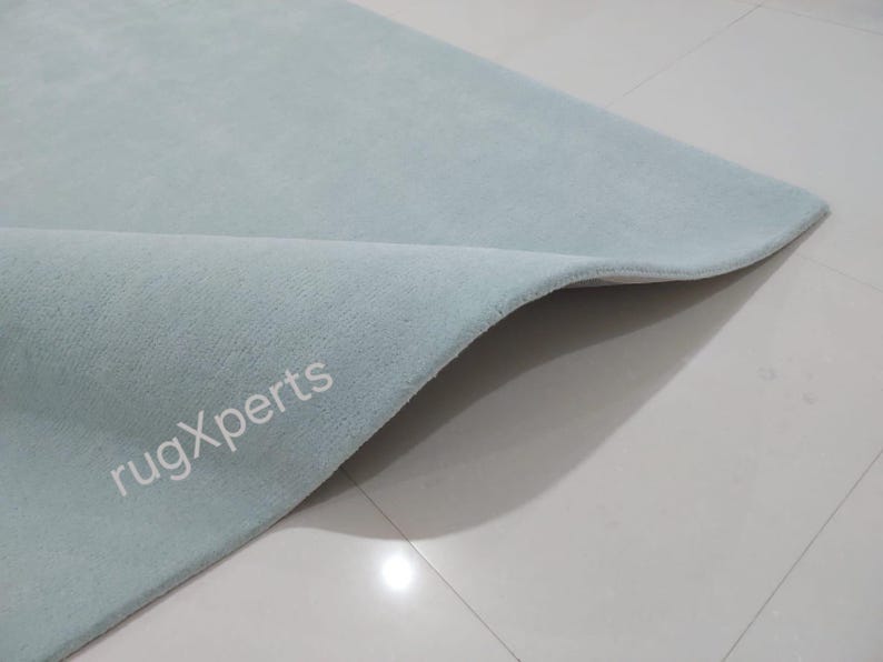 May include: A light blue rug with a soft, textured surface. The corner of the rug is folded over, revealing the underside. The text "rugXperts" is visible on the rug's surface.