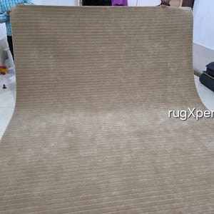 Op de afbeelding: Een groot, beige tapijt met een geribbelde textuur. Het tapijt is uitgerold en toont de lengte en het patroon. De foto is binnenshuis genomen, met een glimp van een persoon en een muur op de achtergrond.