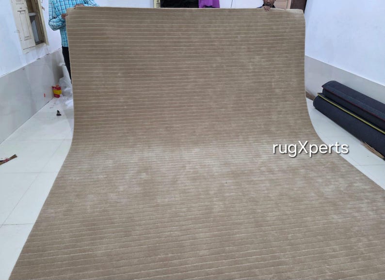 Op de afbeelding: Een groot, beige tapijt met een geribbelde textuur. Het tapijt is uitgerold en toont de lengte en het patroon. De foto is binnenshuis genomen, met een glimp van een persoon en een muur op de achtergrond.