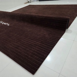 Op de afbeelding: Een opgerolde, rechthoekige bruine vloerkleed met een geribbelde textuur. De vloerkleed is gedeeltelijk uitgerold, waardoor de volledige grootte zichtbaar is. De merknaam "rugXperts" is zichtbaar in witte tekst. De vloerkleed ligt op een witte tegelvloer.