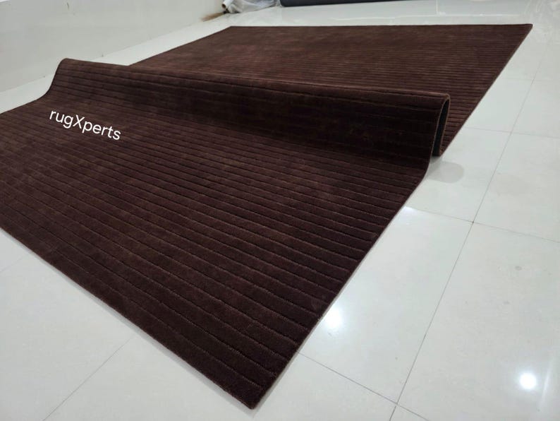 Op de afbeelding: Een opgerolde, rechthoekige bruine vloerkleed met een geribbelde textuur. De vloerkleed is gedeeltelijk uitgerold, waardoor de volledige grootte zichtbaar is. De merknaam "rugXperts" is zichtbaar in witte tekst. De vloerkleed ligt op een witte tegelvloer.