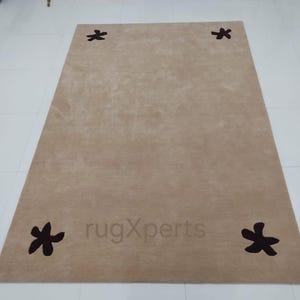 May include: A rectangular beige rug with four dark brown floral designs at each corner. The rug has a soft, textured appearance and is placed on a white tiled floor. The text "rugXperts" is visible on the rug.