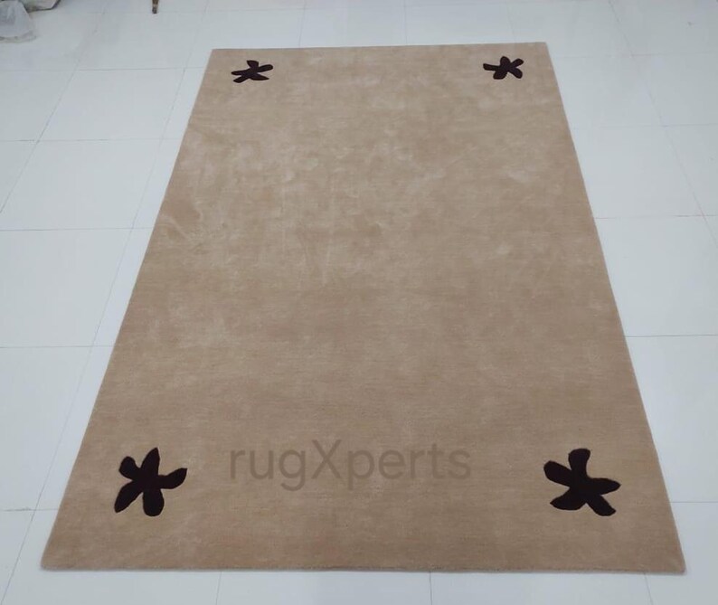 May include: A rectangular beige rug with four dark brown floral designs at each corner. The rug has a soft, textured appearance and is placed on a white tiled floor. The text "rugXperts" is visible on the rug.