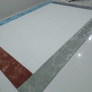 May include: A rectangular rug with a cream-coloured centre and borders in shades of blue, grey, and rust. The text "rugXperts" is visible on the grey border. The rug is placed on a white tiled floor.