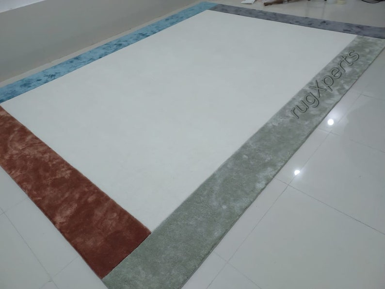 May include: A rectangular rug with a cream-coloured centre and borders in shades of blue, grey, and rust. The text "rugXperts" is visible on the grey border. The rug is placed on a white tiled floor.