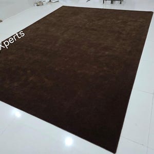 May include: A large, rectangular brown rug. The rug is a solid color and appears to be made of a soft material. The words "rugXperts" are visible in the upper left corner. The rug is placed on a white tiled floor.