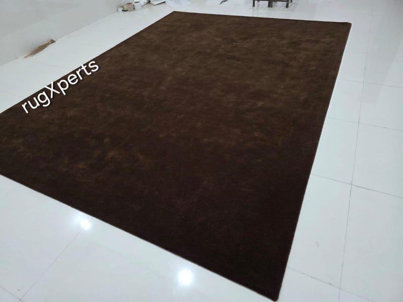 May include: A large, rectangular brown rug. The rug is a solid color and appears to be made of a soft material. The words "rugXperts" are visible in the upper left corner. The rug is placed on a white tiled floor.